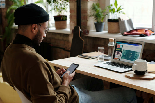 Man in a beret and glasses seated at a wooden table using a smartphone with his laptop open displaying a web page, in a cozy cafe setting displaying home essential gadgets