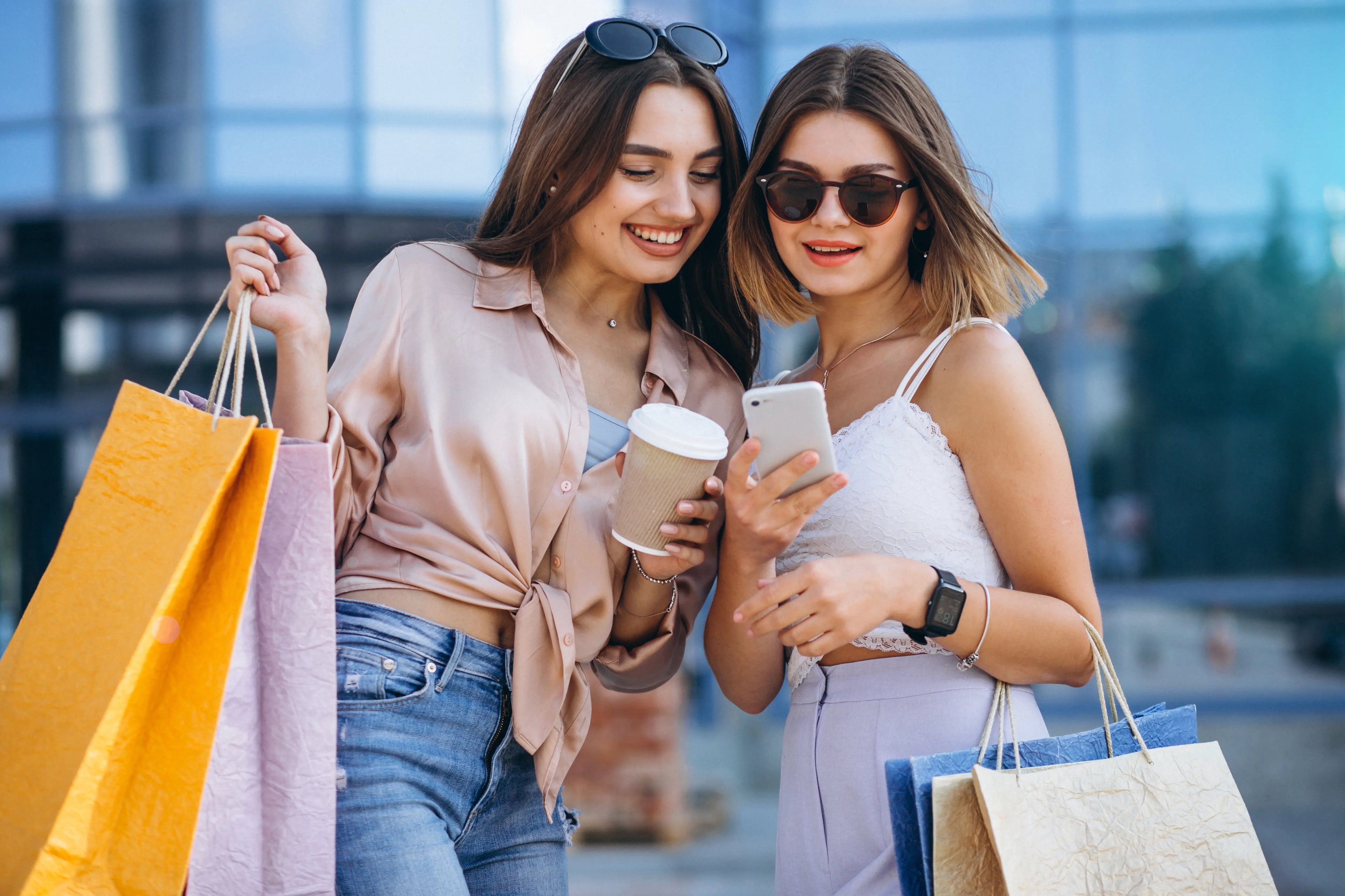 Two women with shopping bags and sunglasses looking at a phone outdoors.